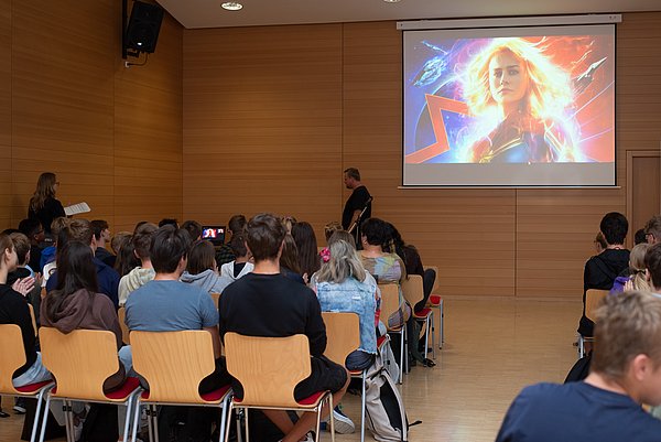 Blick von hinten in einen Vorlesungsraum. Die Zuhörenden sitzen in Stuhlreihen mit einem Mittelgang und sind von hinten zu sehen. Eine Leinwand mit einer Power-Point-Präsentation ist zu sehen und der Referent steht im vorderen Bereich des Raumes.