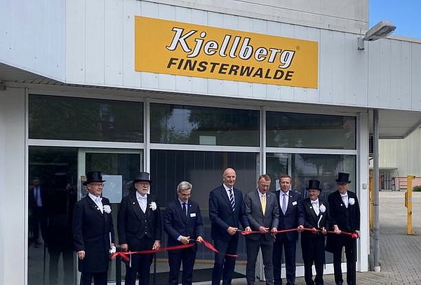 Vor dem Eingang eines Gebäudes mit dem Firmenschild "Kjellberg Finsterwalde" stehen acht Männer in Anzügen und festlicher Kleidung. In der Mitte befindet sich Politiker Dietmar Woidke. Die Männer halten ein rotes Band und zerschneiden dieses symbolisch.