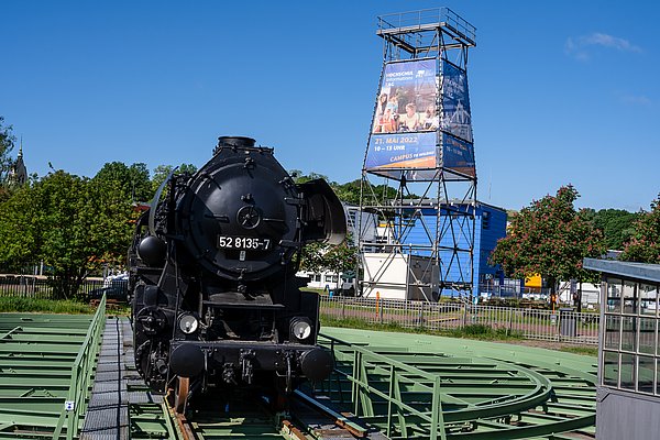 Campusanischt der Technischen Hochschule Wildau mit Blick auf eine Diesellok und Schienensystem. Im Hintergrund steht ein Metallturm mit Bannerwerbung für den Hochschulinformationstag