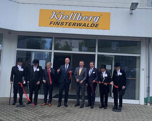 Vor dem Eingang eines Gebäudes mit dem Firmenschild "Kjellberg Finsterwalde" stehen acht Männer in Anzügen und festlicher Kleidung. In der Mitte befindet sich Politiker Dietmar Woidke. Die Männer halten ein rotes Band und zerschneiden dieses symbolisch. 