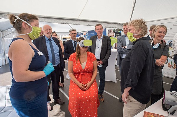 Wissenschaftsministerin des Landes Brandenburg Dr. Manja Schüle sitztend mit VR-Brille in einem Ausstellungspavillion zur Eröffnung der Präsenzstelle Westlausitz Finsterwalde. Um sie herum steht die Delegation der Eröffnungsfrier und die Betreuerin des Standes.