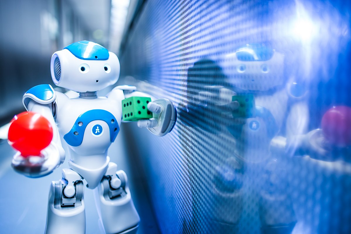 Ein kleiner Roboter hält eine rote Kugel in der rechten Hand und einen grünen Würfel in der linken Hand.