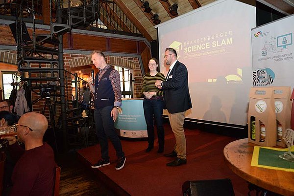 Blick auf die Bühne beim Brandenburger Science Slam 2022 in Finsterwalde. Der Moderator hält ein Mikro in der Hand und eht auf das Publikum zu. Das Orgateam der Präsenzstelle Prignitz steht auf der Bühne.