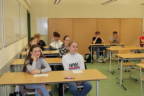 Blick in einen Klassenraum. An den zweier Tischen sitzen Schülerinnen und Schüler und lächelnd in die Kamer oder richten den Blick an der Kamera vorbei in Richtung Lehrkörper, der nicht mit aufgenommen wurde.