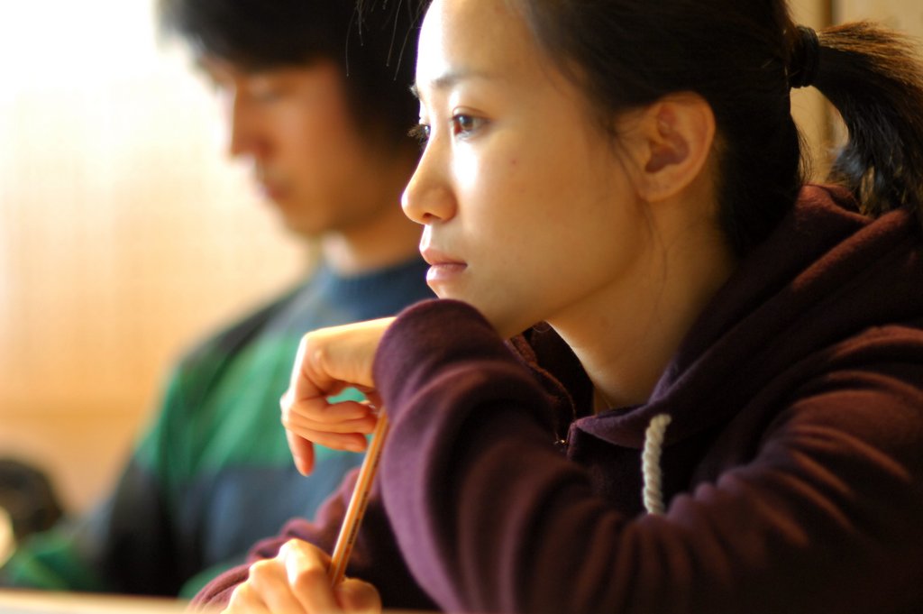 Eine sitzende Studierende lehnt ihre Hand und den Kopf auf einen Stift, den sie mit der zweiten Hand festhält. Neben ihr sitzt ein Kommilitone, der nur verschwommen dargestellt ist.